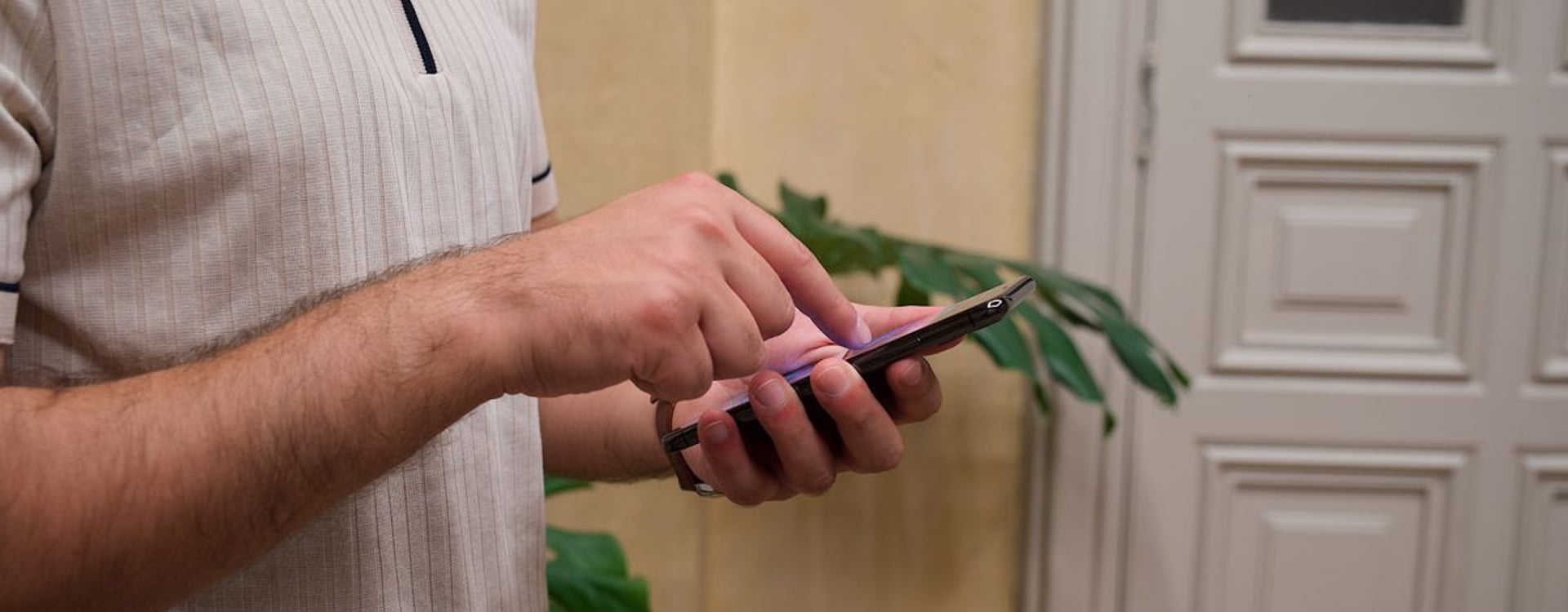 Close-up of a man using smartphone indoors, standing next to a door and plants.