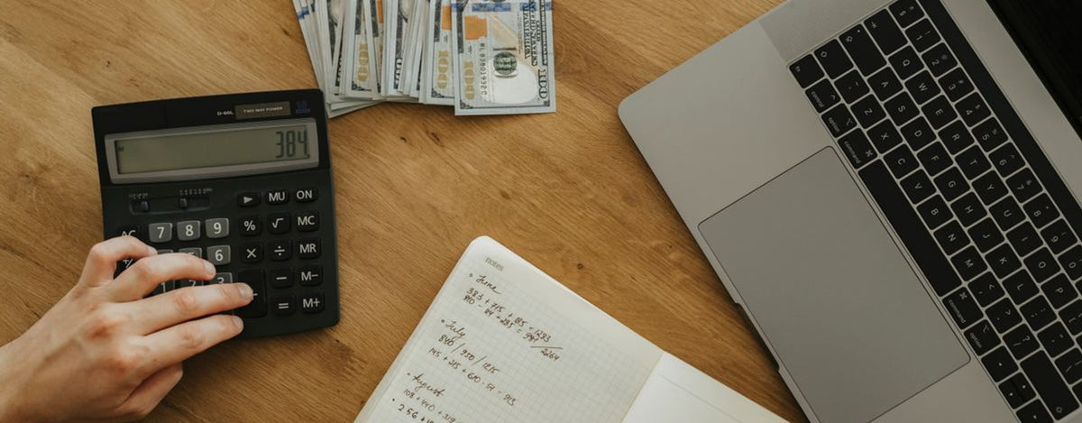 Hands using calculator and notebook, planning finances, with cash and laptop on wooden table.