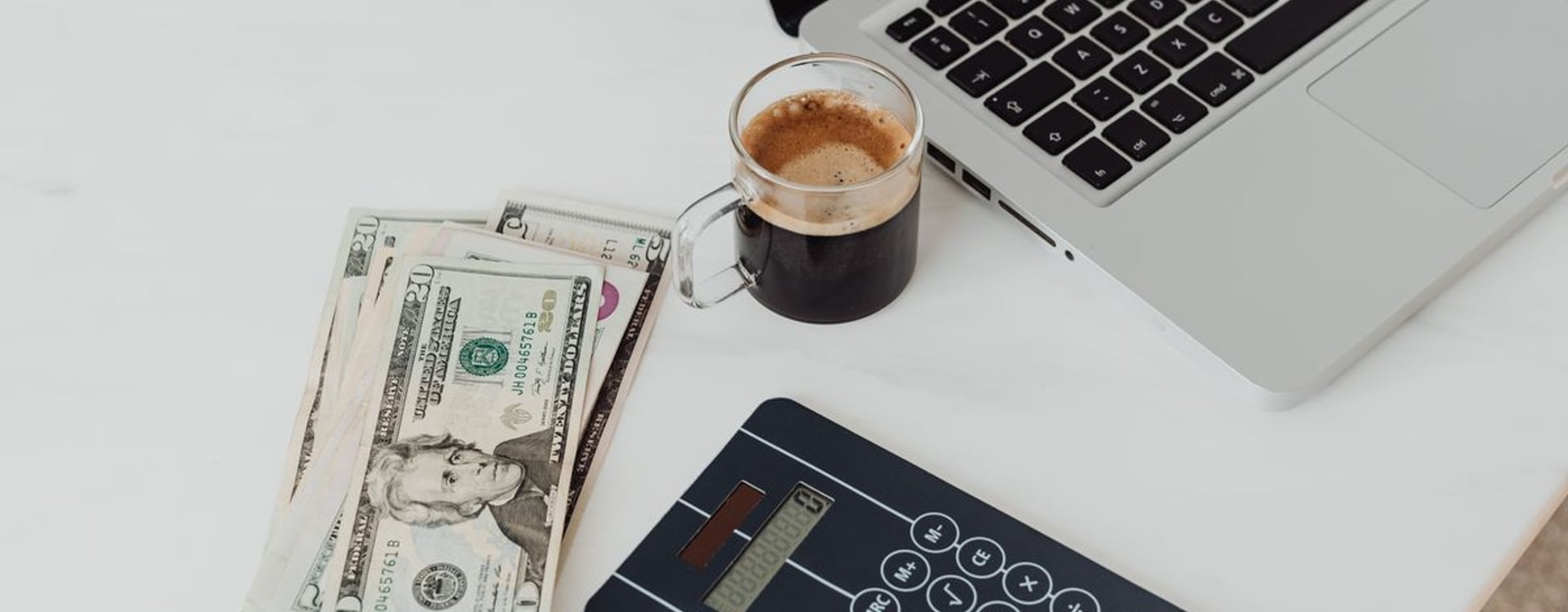 A modern workspace featuring a calculator, cash, and laptop with coffee.